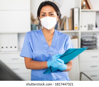 Hispanic Woman General Practitioner In Blue Uniform And Face Mask Standing In Clinic, Filling Out Clipboard With Medical Records