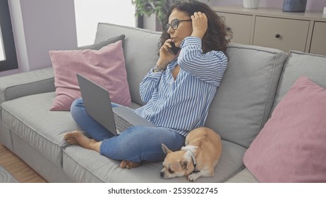 Hispanic woman multitasking with laptop and phone while relaxing with her chihuahua in a cozy living room. - Powered by Shutterstock