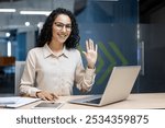 Hispanic woman in office waving at camera, seated at desk with laptop, phone, documents. Reflects friendly work environment, professional interaction, positive communication, confident demeanor.