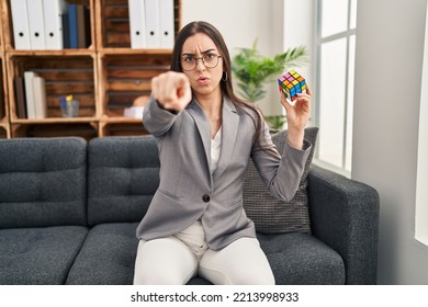 Hispanic Woman Playing Colorful Puzzle Cube Intelligence Game Pointing With Finger To The Camera And To You, Confident Gesture Looking Serious 