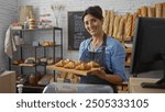 Hispanic woman proudly presenting fresh croissants in a bakery, showcasing a variety of baked goods on shelves in a cozy indoor shop setting.