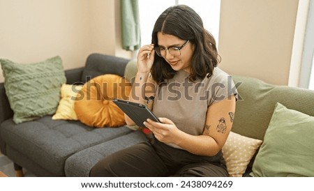 Hispanic woman reading tablet on couch in cozy living room setting, expressing comfort and leisure.