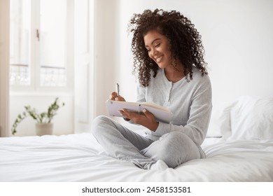 A Hispanic woman is seated on a bed, focused on writing in a notebook. She appears engrossed in her writing, pen in hand, surrounded by a cozy bedroom setting. - Powered by Shutterstock