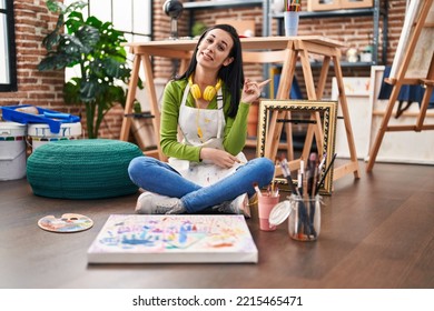 Hispanic Woman Sitting At Art Studio Painting On Canvas Smiling Happy Pointing With Hand And Finger To The Side 