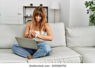 Hispanic Woman Sitting On The Sofa At Home Using Laptop Checking The Time On Wrist Watch, Relaxed And Confident 