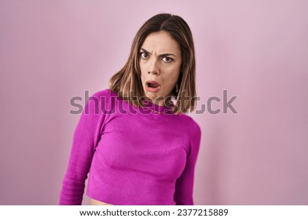 Hispanic woman standing over pink background in shock face, looking skeptical and sarcastic, surprised with open mouth 
