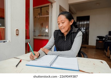 Hispanic woman studying at home - Happy senior adult writing in a notebook - Retired adult doing budgeting at home - Mayan adult woman learning at home - Powered by Shutterstock