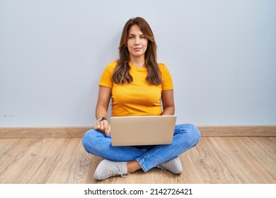 Hispanic Woman Using Laptop Sitting On The Floor At Home Relaxed With Serious Expression On Face. Simple And Natural Looking At The Camera. 