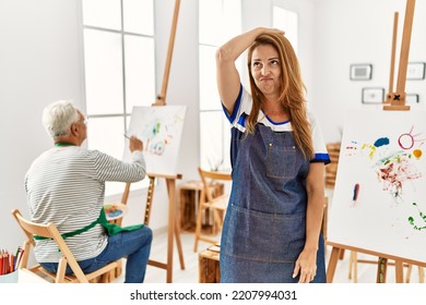 Hispanic woman wearing apron at art studio confuse and wonder about question. uncertain with doubt, thinking with hand on head. pensive concept. 