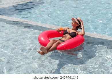 Hispanic woman, wearing swimming costume, hat sunglasses, floats on float placed on surface water, woman holding glass water in hand, drinking water while sitting in swimming pool, sunbathing. - Powered by Shutterstock