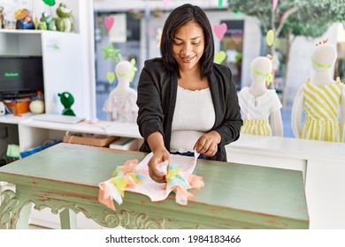 Hispanic woman working as shop assistant at children clothes small retail trade. sales assistant smiling happy at shopping counter while folding shirt.