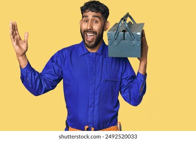 Hispanic worker with toolbox in studio receiving a pleasant surprise, excited and raising hands.