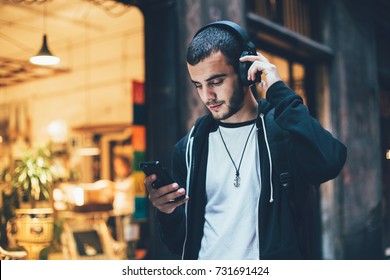 Hispanic young attractive man stands in dark street in front of shop, changes songs and tracks on smartphone, listens to music in wireless headphones. Hipster with slight beard - Shutterstock ID 731691424