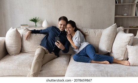 Hispanic Young Couple Relaxing On Cozy Couch Looking At Cell Phone Screen Laughing Watch Funny Online Videos, Amusing Internet Content, Have Fun Use Modern Wireless Tech Spend Carefree Weekend At Home