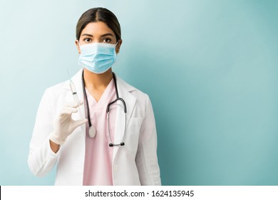 Hispanic Young Doctor Holding Syringe While Wearing Mask Against Blue Background