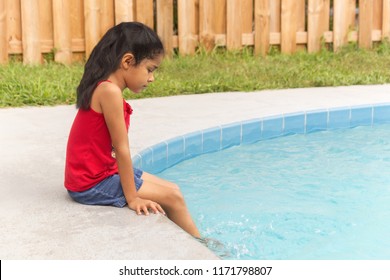 A Hispanic Young Girl Sits Peacefully By The Edge Of The Pool Dangling Her Feet In The Pool.