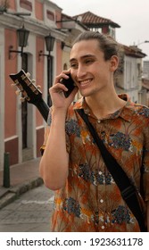 Hispanic Young Man In The Center Of Bogota Colombia, La Candelaria, A Place Of Colonial Houses Perfect For A Tourist Destination, Talking On A Cell Phone, Holding A Guitar