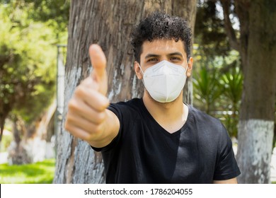 Hispanic Young Man With Disposable Mask And Thumb Up - Young Man In The Park Sitting Under A Tree On A Sunny Day With Preventive Measures To Avoid Catching Any Virus - Social Distancing