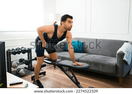 Hispanic young man in his 20s doing bench row exercises using weights while training in the living room