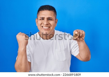 Hispanic young man holding virtual currency bitcoin screaming proud, celebrating victory and success very excited with raised arms 