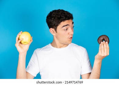 Hispanic young man looking at doughnut and holding an apple in the other hand isolated on blue background.