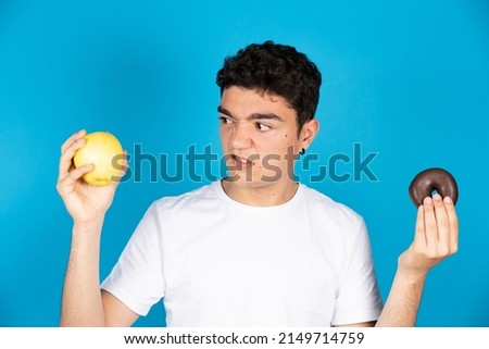Hispanic young man looking with repugnance at apple while holding a chocolate donut or doughnut in the other hand isolated on blue background