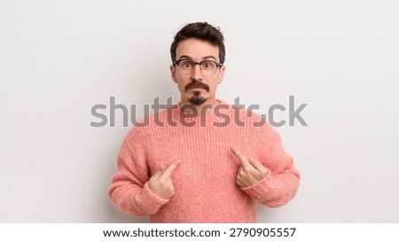 hispanic young man pointing to self with a confused and quizzical look, shocked and surprised to be chosen