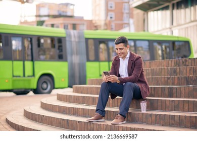 Hispanic Young Man Sitting In A City Square With His Cell Phone And A Public Bus In The Background.