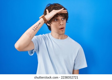 Hispanic young man standing over blue background very happy and smiling looking far away with hand over head. searching concept. 