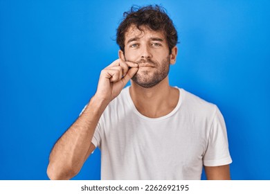 Hispanic young man standing over blue background mouth and lips shut as zip with fingers. secret and silent, taboo talking 
