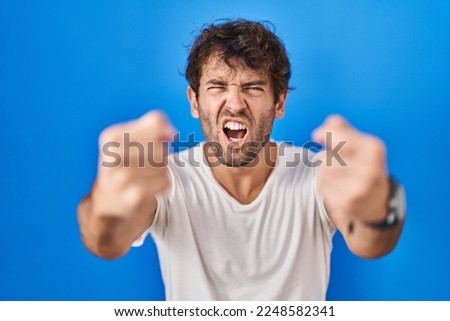 Hispanic young man standing over blue background angry and mad raising fists frustrated and furious while shouting with anger. rage and aggressive concept. 