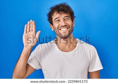Hispanic young man standing over blue background waiving saying hello happy and smiling, friendly welcome gesture 