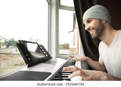 Hispanic young man with a stubble wearing a grey hat plays piano and smiles while following an online piano lesson on his tablet sitting next to a bay window