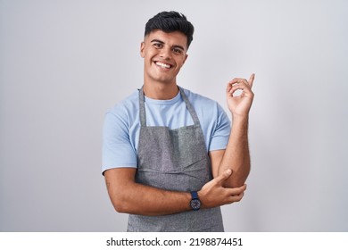 Hispanic Young Man Wearing Apron Over White Background With A Big Smile On Face, Pointing With Hand Finger To The Side Looking At The Camera. 