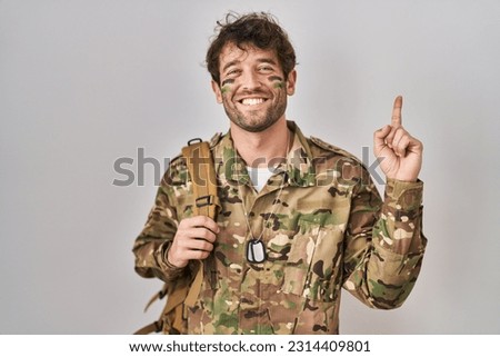 Hispanic young man wearing camouflage army uniform with a big smile on face, pointing with hand finger to the side looking at the camera. 