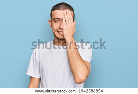 Hispanic young man wearing casual white t shirt covering one eye with hand, confident smile on face and surprise emotion. 
