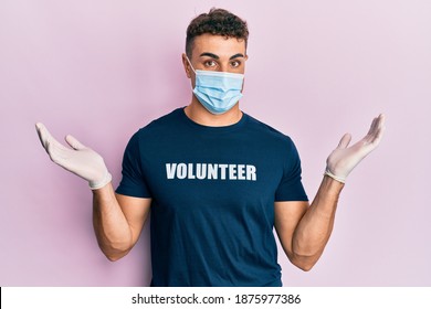 Hispanic Young Man Wearing Medical Mask And Volunteer T Shirt Clueless And Confused With Open Arms, No Idea And Doubtful Face. 