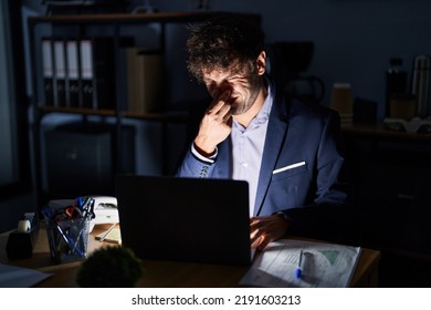 Hispanic Young Man Working At The Office At Night Smelling Something Stinky And Disgusting, Intolerable Smell, Holding Breath With Fingers On Nose. Bad Smell 