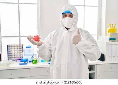 Hispanic Young Man Working At Scientist Laboratory Holding Virus Toy Smiling Happy And Positive, Thumb Up Doing Excellent And Approval Sign 