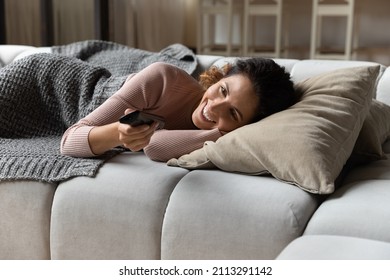 Hispanic young smiling woman lying on couch cushion holds TV remote controller switch channels watch internet television, single 30s female enjoy day off at cozy home. Lifestyle, lazy holidays concept