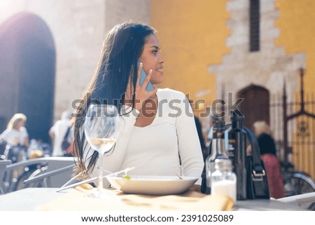 Hispanic young woman eating alone in cafe patio, talking by phone, waiting for someone during sunny weather in European cafeteria. Communication, meeting, dating, chatting with friends.