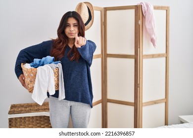 Hispanic Young Woman Holding Laundry Basket Pointing With Finger To The Camera And To You, Confident Gesture Looking Serious 