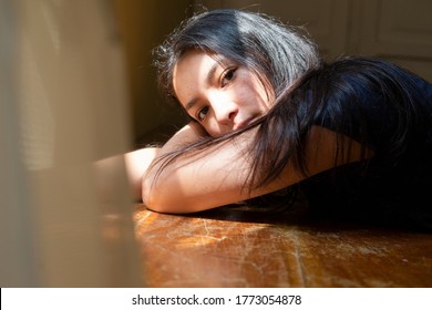 Hispanic Young Woman Lying On Her Arms At A Table With Sun Rays On Her Face, Young Woman Thinking At Home Enjoying The Sun, Sad And Serious Woman, Looking At The Camera.