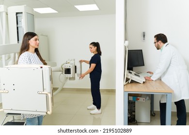 Hispanic Young Woman At The Medical Imaging Lab Getting An X-ray Test Of Her Back And Chest With The Help Of A Technician And Radiologist 