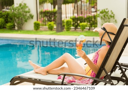 Hispanic young woman relaxing in the hotel lounger enjoying drinking a cocktail in her swimsuit by the pool outdoors