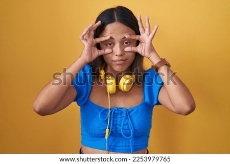Hispanic young woman standing over yellow background trying to open eyes with fingers, sleepy and tired for morning fatigue 
