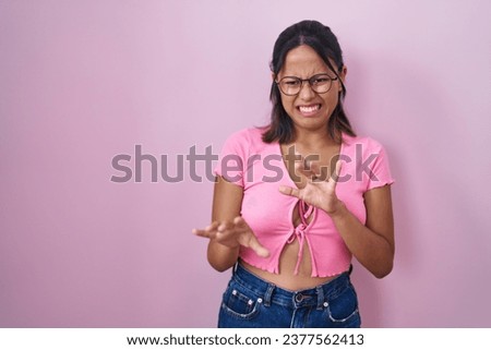 Hispanic young woman standing over pink background wearing glasses disgusted expression, displeased and fearful doing disgust face because aversion reaction. with hands raised 