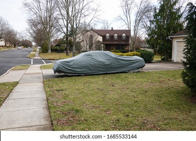 Historic Antique Muscle Car In The Driveway Of A Suburban House Completely Covered With A Gray Car Protection Cover