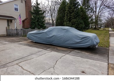 Historic Antique Muscle Car In The Driveway Of A Suburban House Completely Covered With A Gray Car Protection Cover