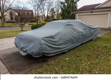 Historic Antique Muscle Car In The Driveway Of A Suburban House Completely Covered With A Gray Car Protection Cover